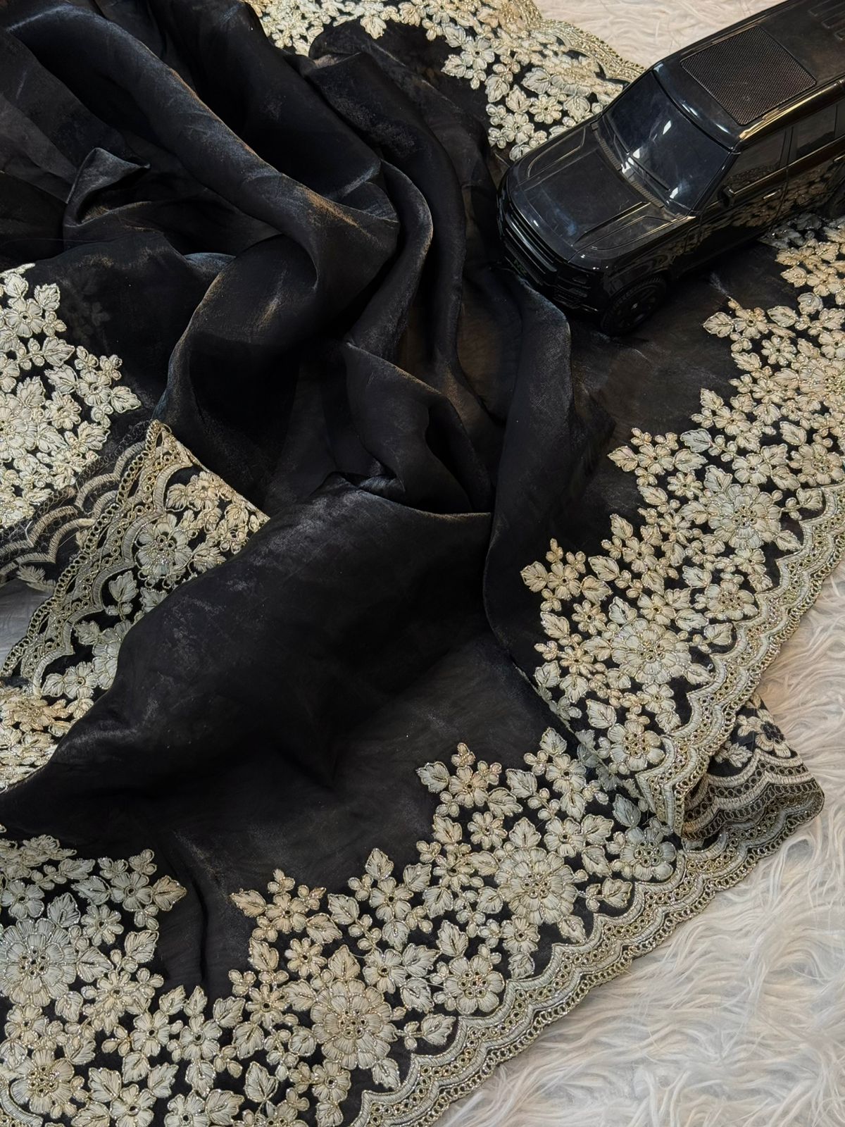 A classy black Fandy silk saree decorated with elegant white floral embroidery and a beautifully scalloped border. The matching blouse completes this stylish and graceful traditional ensemble.