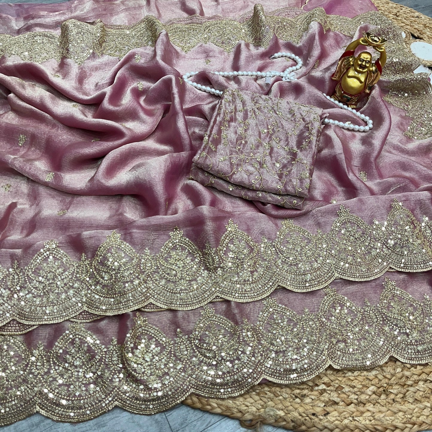 A blush pink saree decorated with golden threadwork and intricate sequin patterns along a wide scalloped border. The matching blouse piece complements the saree’s luxurious and royal appeal.