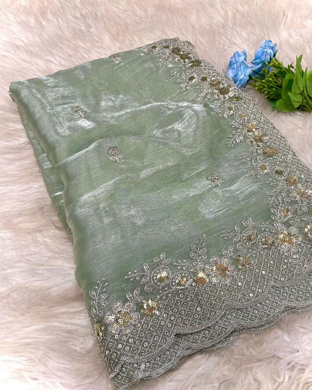 A mint green organza saree with an elegant shimmer, featuring intricate silver and gold sequin embroidery along the scalloped border. The saree is paired with a contrasting olive green blouse piece, placed in a woven basket on a white fur background.