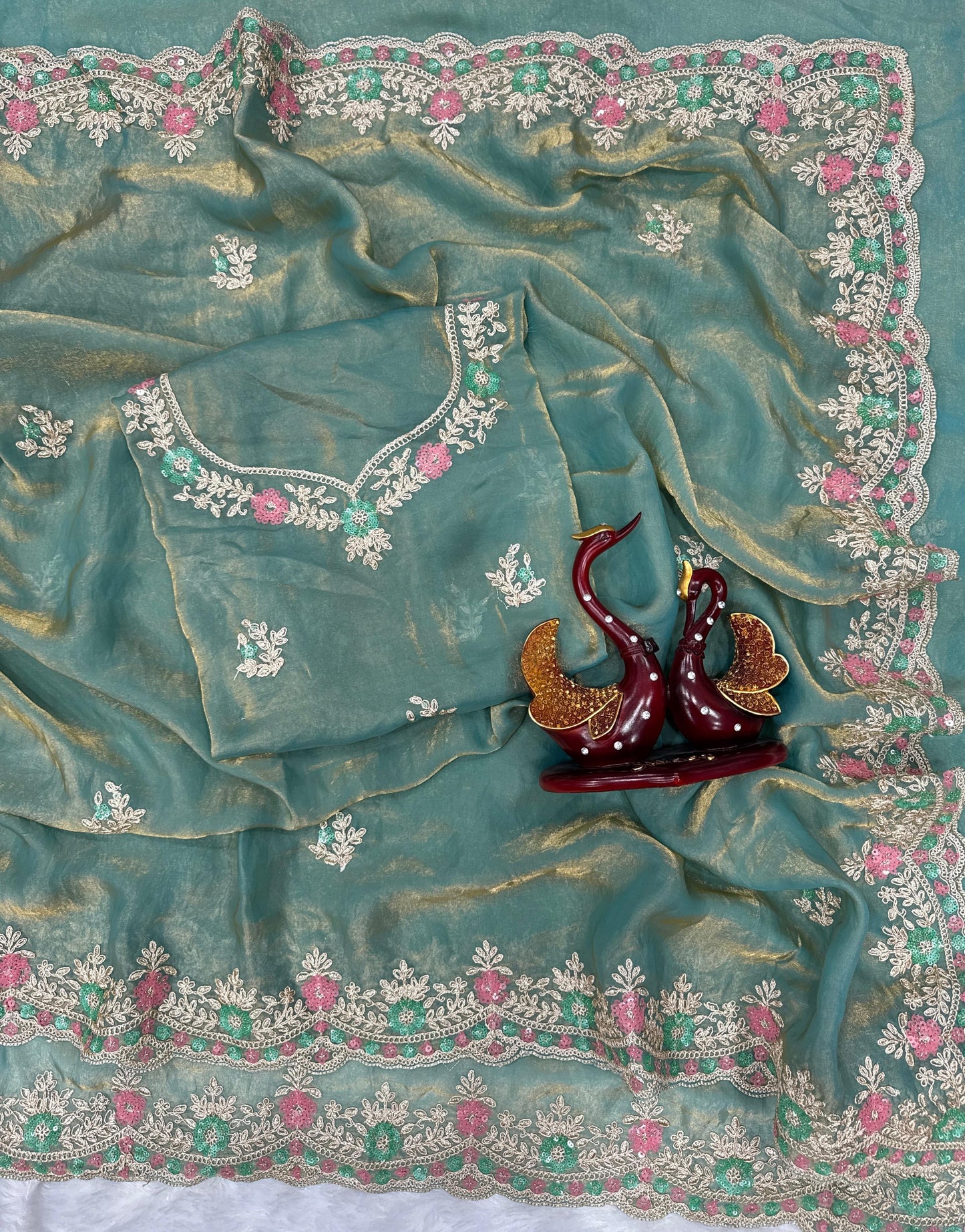 A woman wearing a space blue silk saree featuring a delicate pink and silver floral embroidered border. The saree has a soft, glossy finish paired with a matching embroidered blouse. She accessorizes with a pearl choker, earrings, and statement jewelry, posing gracefully on a balcony with greenery in the background, showcasing the saree’s elegant drape and intricate detailing.