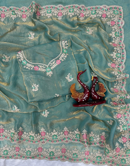 A woman wearing a space blue silk saree featuring a delicate pink and silver floral embroidered border. The saree has a soft, glossy finish paired with a matching embroidered blouse. She accessorizes with a pearl choker, earrings, and statement jewelry, posing gracefully on a balcony with greenery in the background, showcasing the saree’s elegant drape and intricate detailing.