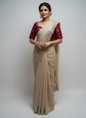 A beige gold saree with intricate zari embroidery along the border, paired with a rich wine-red embroidered blouse. The elegant color contrast and luxurious texture make it perfect for festive and wedding occasions.