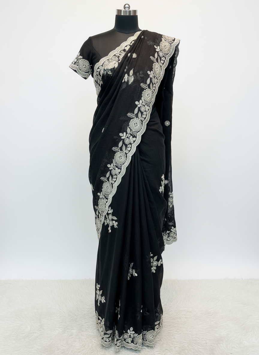 A luxurious black saree featuring intricate silver floral embroidery and a scalloped lace border. The saree is displayed with a matching embroidered blouse piece, laid on a soft fur background, highlighting its elegant craftsmanship and rich fabric texture.