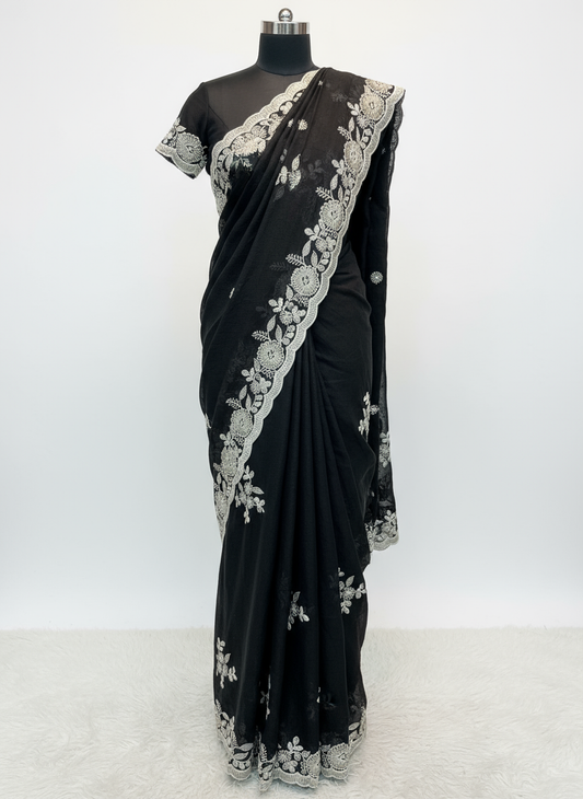 A luxurious black saree featuring intricate silver floral embroidery and a scalloped lace border. The saree is displayed with a matching embroidered blouse piece, laid on a soft fur background, highlighting its elegant craftsmanship and rich fabric texture.