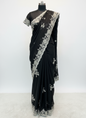 A luxurious black saree featuring intricate silver floral embroidery and a scalloped lace border. The saree is displayed with a matching embroidered blouse piece, laid on a soft fur background, highlighting its elegant craftsmanship and rich fabric texture.