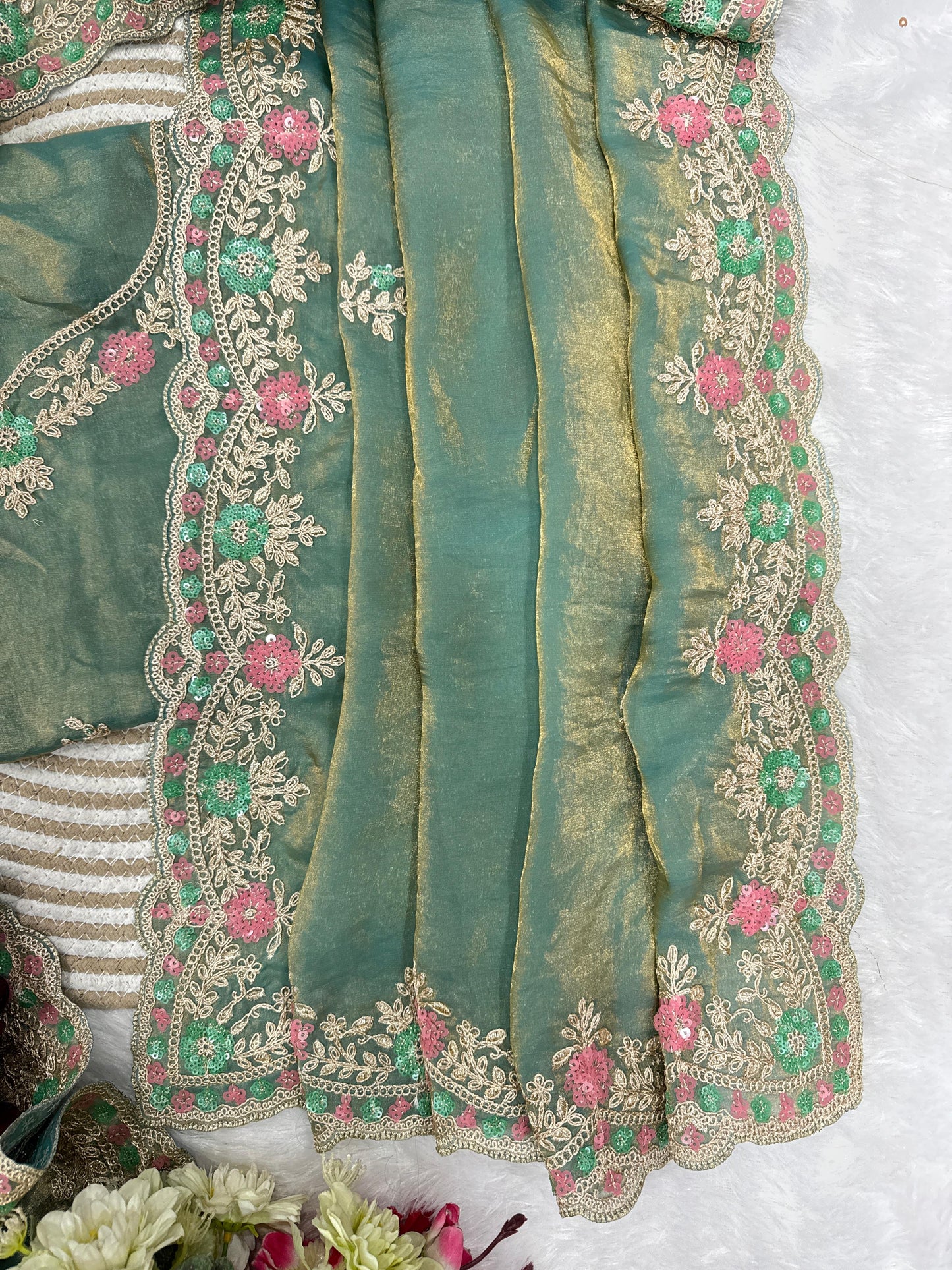 A woman elegantly draped in an aqua space silk saree featuring a shimmering hand-embroidered floral border in soft pink and silver tones. She pairs it with a matching embroidered blouse and pearl jewelry, posing gracefully against a lush green outdoor backdrop.