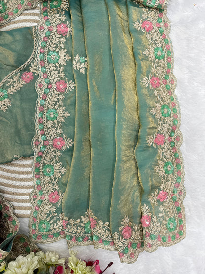 A woman elegantly draped in an aqua space silk saree featuring a shimmering hand-embroidered floral border in soft pink and silver tones. She pairs it with a matching embroidered blouse and pearl jewelry, posing gracefully against a lush green outdoor backdrop.
