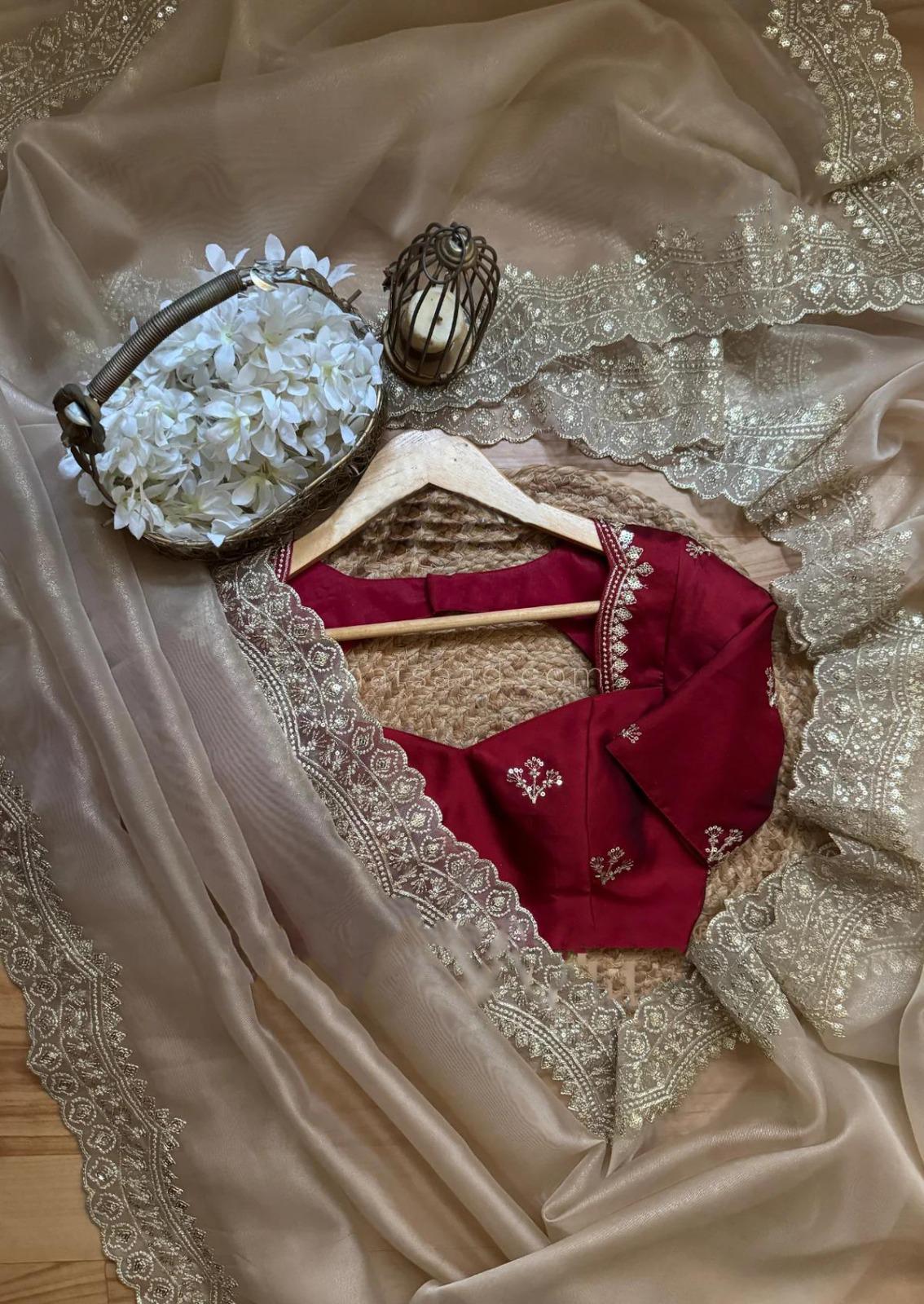 An elegant champagne beige saree featuring a shimmering embroidered border, paired with a deep maroon blouse embellished with golden threadwork. The soft neutral tones with a bold contrast create a sophisticated modern ethnic look.
