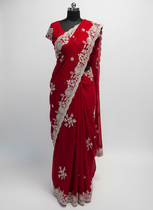 A rich crimson red saree adorned with intricate white floral embroidery and a scalloped lace border. The fabric includes a matching embroidered blouse piece, displayed elegantly on a soft fur surface, highlighting the saree’s vibrant hue and fine craftsmanship.