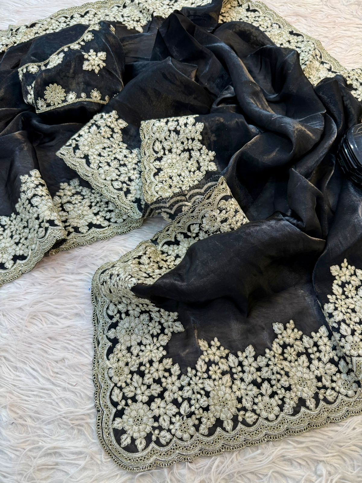 A premium black Fandy silk saree adorned with detailed handcrafted white floral embroidery. The heavy scalloped border and matching embroidered blouse add a sophisticated and festive appeal to the outfit.