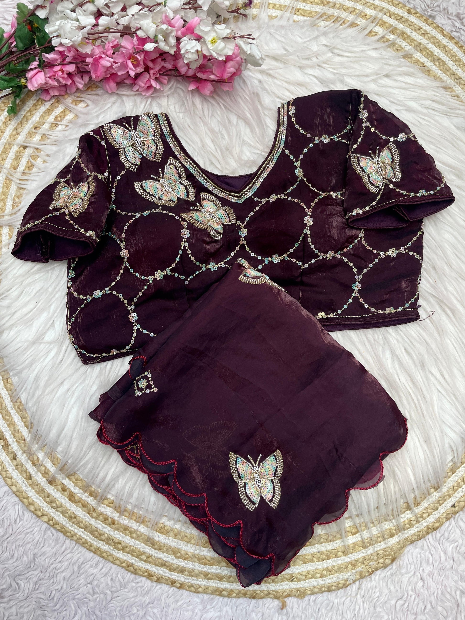 A luxurious maroon space silk saree beautifully detailed with shimmering butterfly embroidery and a scalloped edge. The outfit includes a stitched blouse with matching embroidery and geometric sequin work, ideal for weddings and parties.