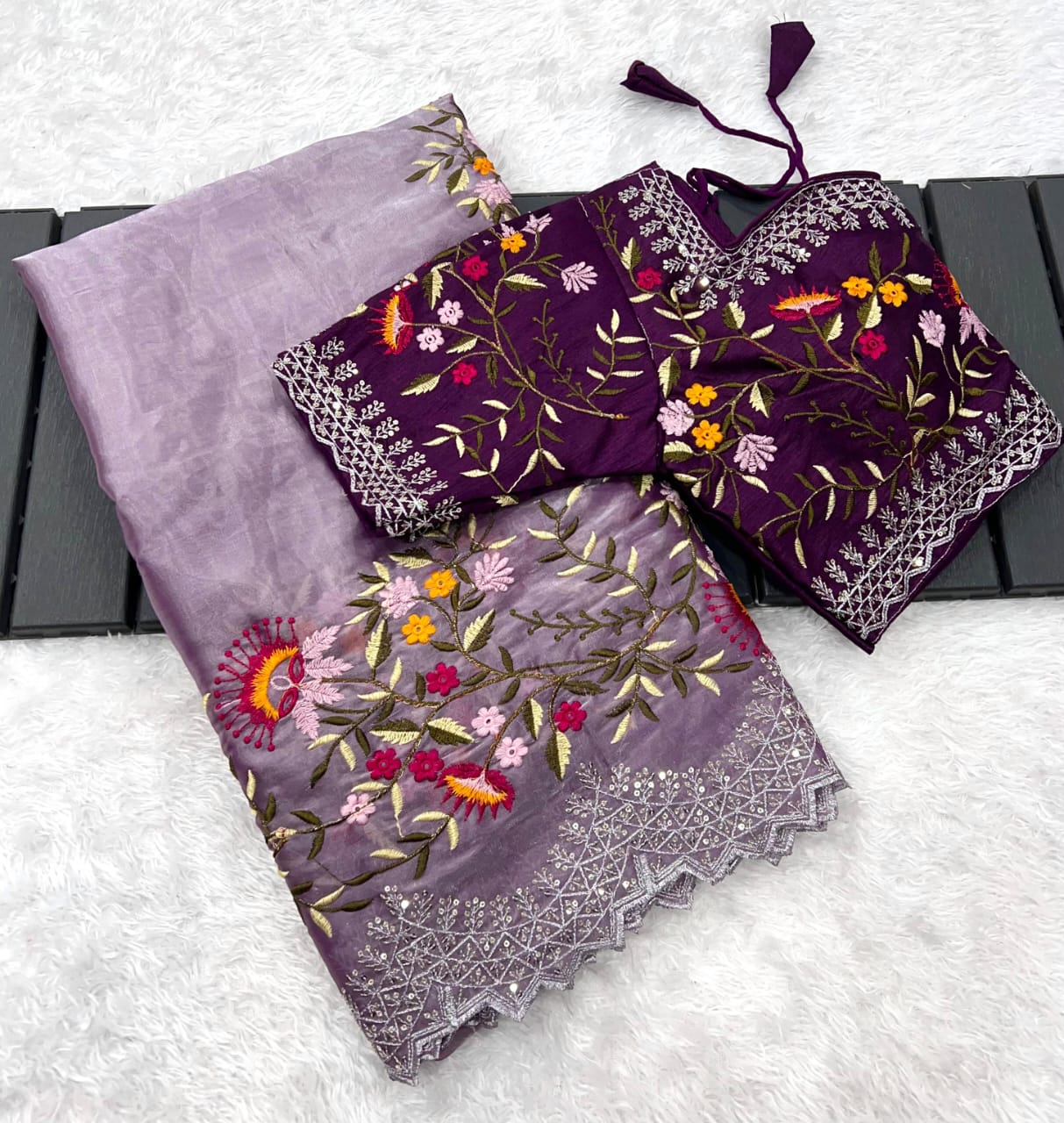 A designer onion shade saree made from soft organza fabric, embellished with colorful floral embroidery and shimmering lace edging. The matching stitched blouse with coordinated detailing adds a rich festive appeal.