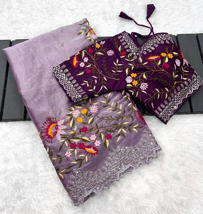 A designer onion shade saree made from soft organza fabric, embellished with colorful floral embroidery and shimmering lace edging. The matching stitched blouse with coordinated detailing adds a rich festive appeal.