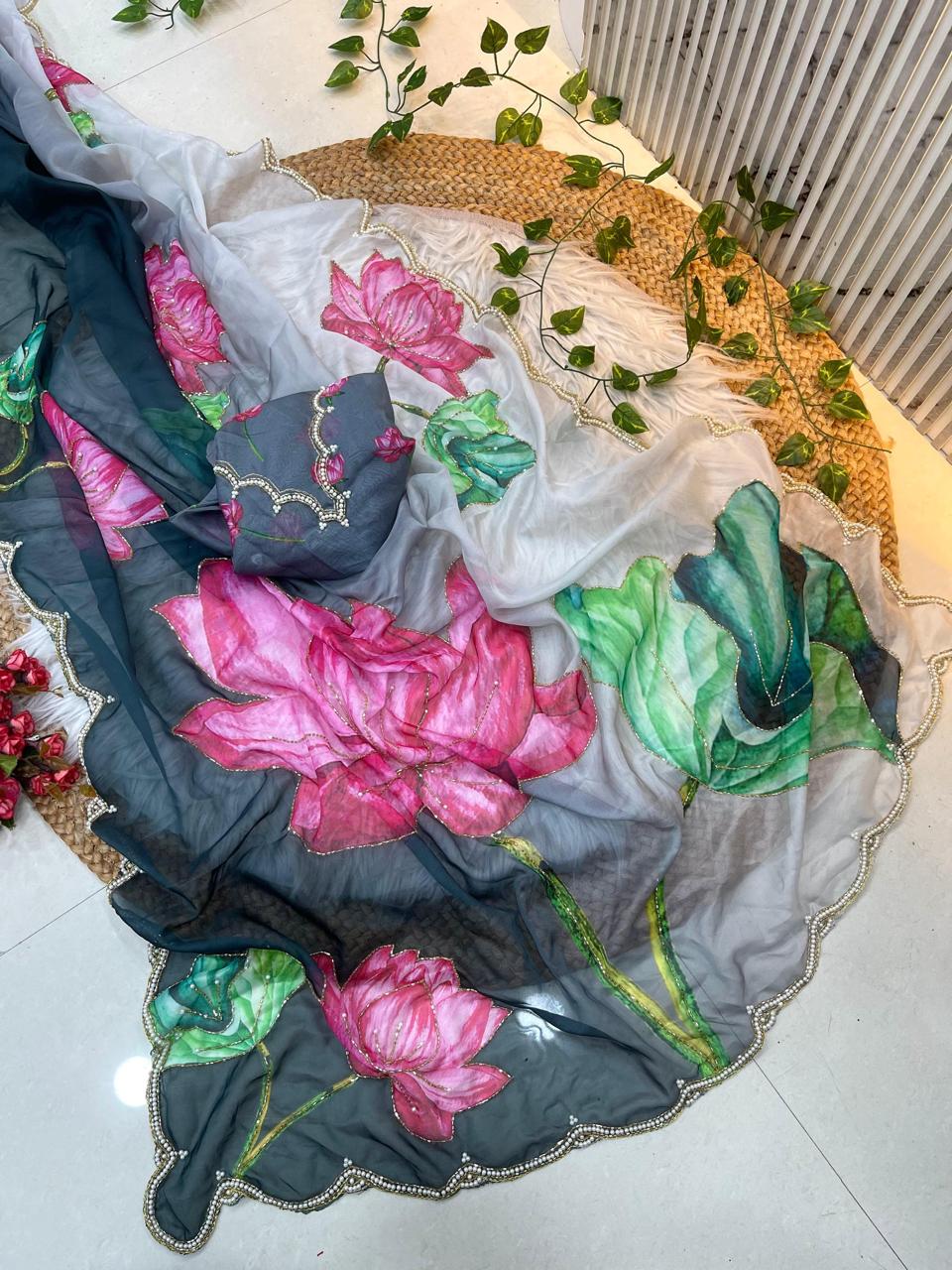 A designer organza saree adorned with watercolor-style lotus flowers in bright pink and lush green tones. The saree includes a scalloped pearl border and a matching lotus-embroidered blouse, offering a rich and elegant festive appearance.
