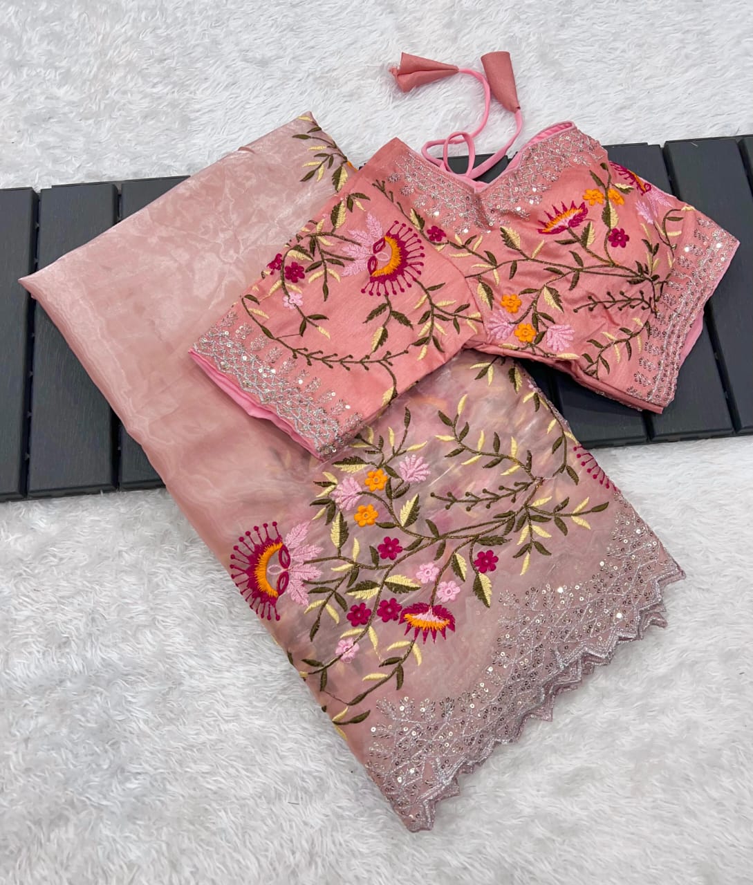A designer peach saree crafted from lightweight organza fabric, adorned with colorful floral embroidery and scalloped lace edging. The stitched blouse features matching embroidery, making this ensemble perfect for festive and celebratory occasions.