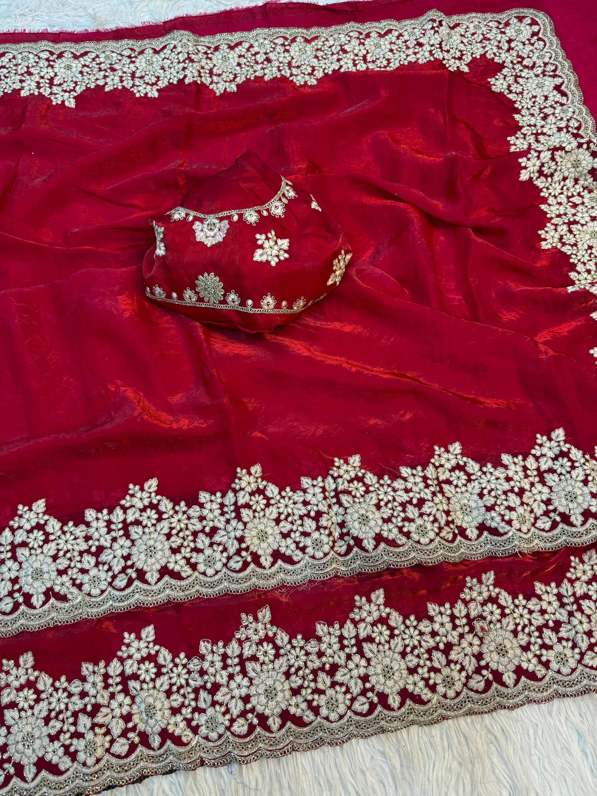 A designer red Fandy silk saree beautifully embellished with handcrafted white floral embroidery on the border. The matching blouse enhances the luxurious and sophisticated appeal of this festive ensemble.