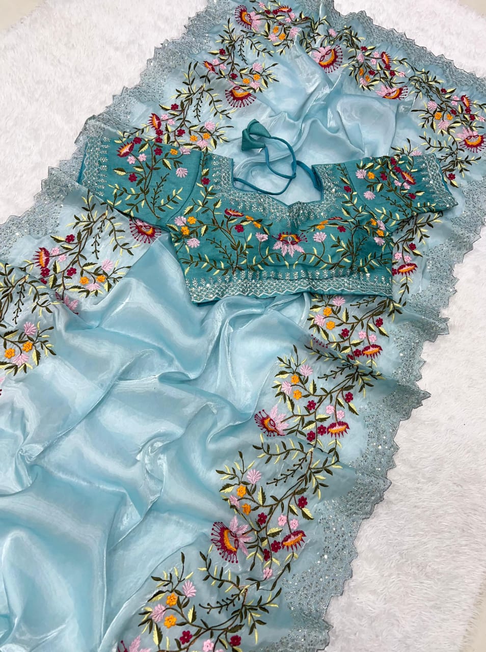 A designer sky blue saree made from sheer organza fabric, embellished with colorful floral embroidery. The set includes a ready-to-wear blouse with matching floral threadwork, blending modern sophistication with traditional beauty.