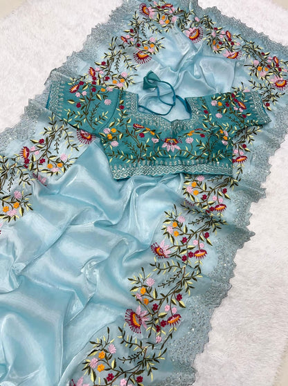 A designer sky blue saree made from sheer organza fabric, embellished with colorful floral embroidery. The set includes a ready-to-wear blouse with matching floral threadwork, blending modern sophistication with traditional beauty.