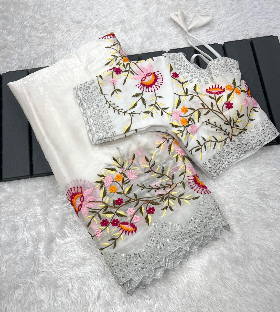 A designer white saree made from lightweight organza fabric, embellished with colorful floral embroidery and lace detailing. The outfit includes a stitched blouse with matching embroidery, perfect for festive occasions and elegant celebrations.