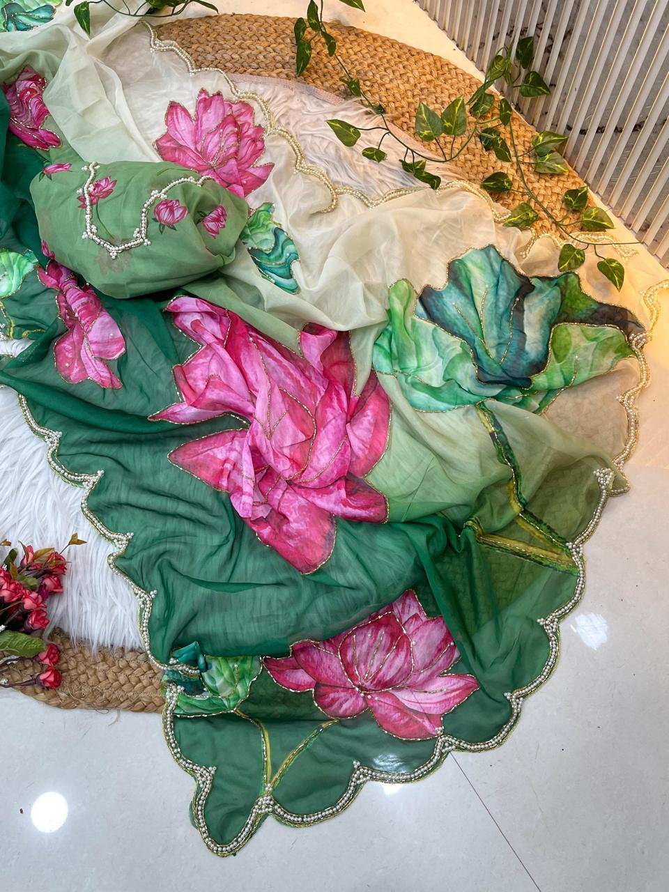 A stylish green organza saree decorated with bold multicolor lotus artwork and soft cream shading. The saree has a delicate pearl scalloped border, paired with a matching embroidered blouse for a festive and fashionable look.