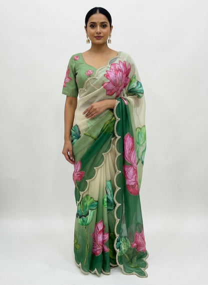 A stunning green soft organza saree featuring large hand-painted pink lotus flowers and vibrant green leaf artwork. The saree has a pearl-embellished scalloped border and comes with a matching blouse showcasing lotus detailing, creating a graceful and artistic ethnic look.