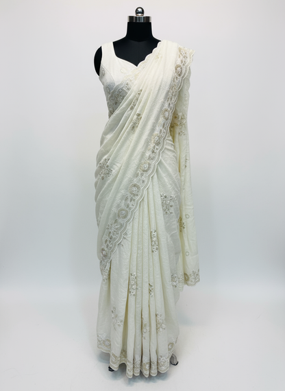 An ivory white saree featuring delicate silver floral embroidery and a scalloped lace border. The saree is paired with a matching embroidered blouse piece, elegantly spread on a soft fur background, highlighting its graceful texture and intricate craftsmanship.