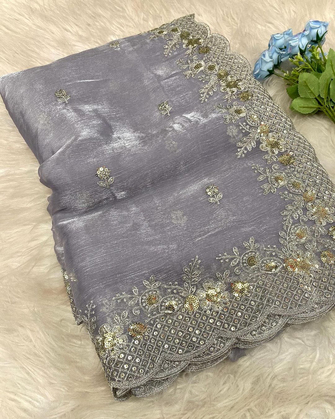 A soft lavender organza saree adorned with intricate silver and gold sequin embroidery along a scalloped border. The saree has a delicate shimmer and is paired with a contrasting deep magenta blouse piece, arranged elegantly in a woven basket on a white fur background.