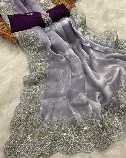 A soft lavender organza saree adorned with intricate silver and gold sequin embroidery along a scalloped border. The saree has a delicate shimmer and is paired with a contrasting deep magenta blouse piece, arranged elegantly in a woven basket on a white fur background.