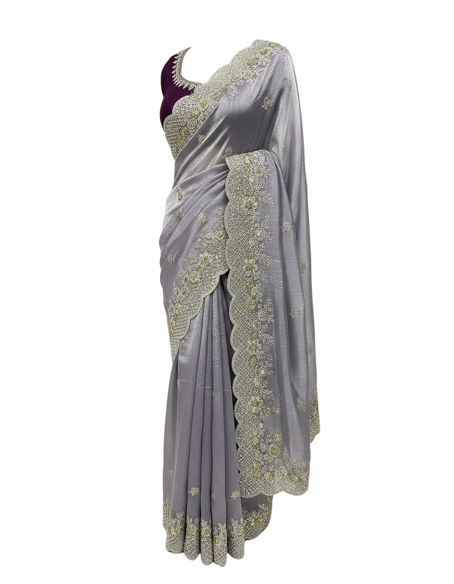 A soft lavender organza saree adorned with intricate silver and gold sequin embroidery along a scalloped border. The saree has a delicate shimmer and is paired with a contrasting deep magenta blouse piece, arranged elegantly in a woven basket on a white fur background.