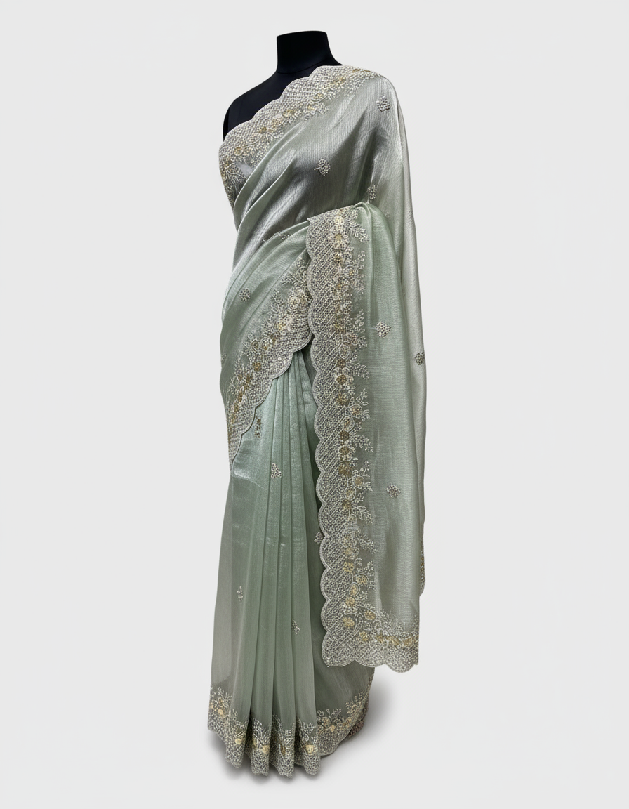 A mint green organza saree with an elegant shimmer, featuring intricate silver and gold sequin embroidery along the scalloped border. The saree is paired with a contrasting olive green blouse piece, placed in a woven basket on a white fur background.
