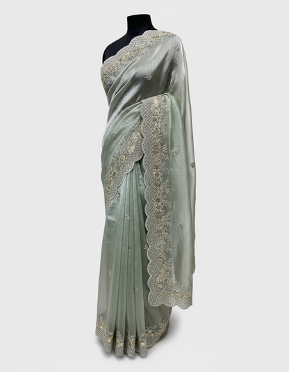 A mint green organza saree with an elegant shimmer, featuring intricate silver and gold sequin embroidery along the scalloped border. The saree is paired with a contrasting olive green blouse piece, placed in a woven basket on a white fur background.
