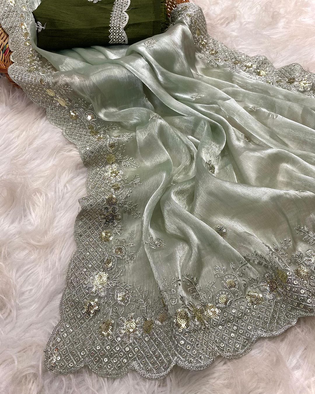 A mint green organza saree with an elegant shimmer, featuring intricate silver and gold sequin embroidery along the scalloped border. The saree is paired with a contrasting olive green blouse piece, placed in a woven basket on a white fur background.