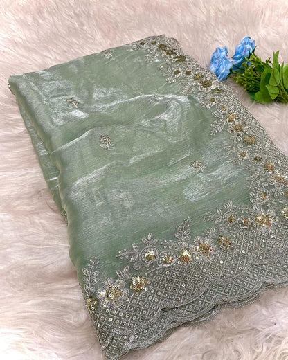 A mint green organza saree with an elegant shimmer, featuring intricate silver and gold sequin embroidery along the scalloped border. The saree is paired with a contrasting olive green blouse piece, placed in a woven basket on a white fur background.