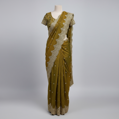 A luxurious mustard gold organza saree adorned with intricate golden embroidery and a richly embellished scalloped border. The saree is paired with a matching embroidered blouse piece, displayed with elegant accessories, showcasing a regal and festive aesthetic.