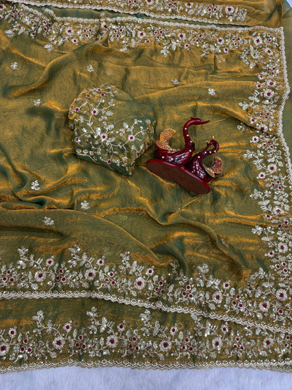 An olive-gold Fandy silk saree detailed with floral embroidery and shimmering zari accents. The saree’s ornate border and soft sheen give it a graceful and elegant appearance suitable for both festive and formal occasions.