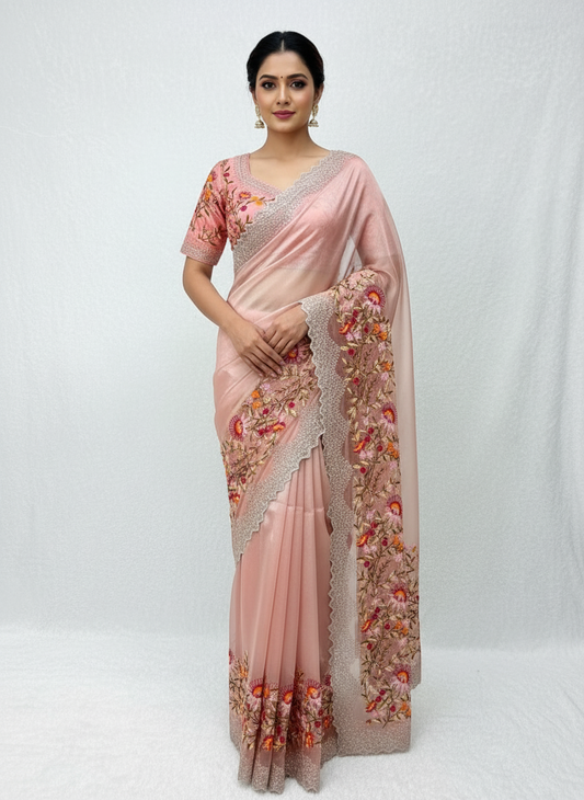 A delicate peach organza saree featuring vibrant multicolor floral embroidery along the border. The saree is paired with a matching stitched blouse showcasing intricate threadwork and sequin detailing, creating a graceful and festive look.