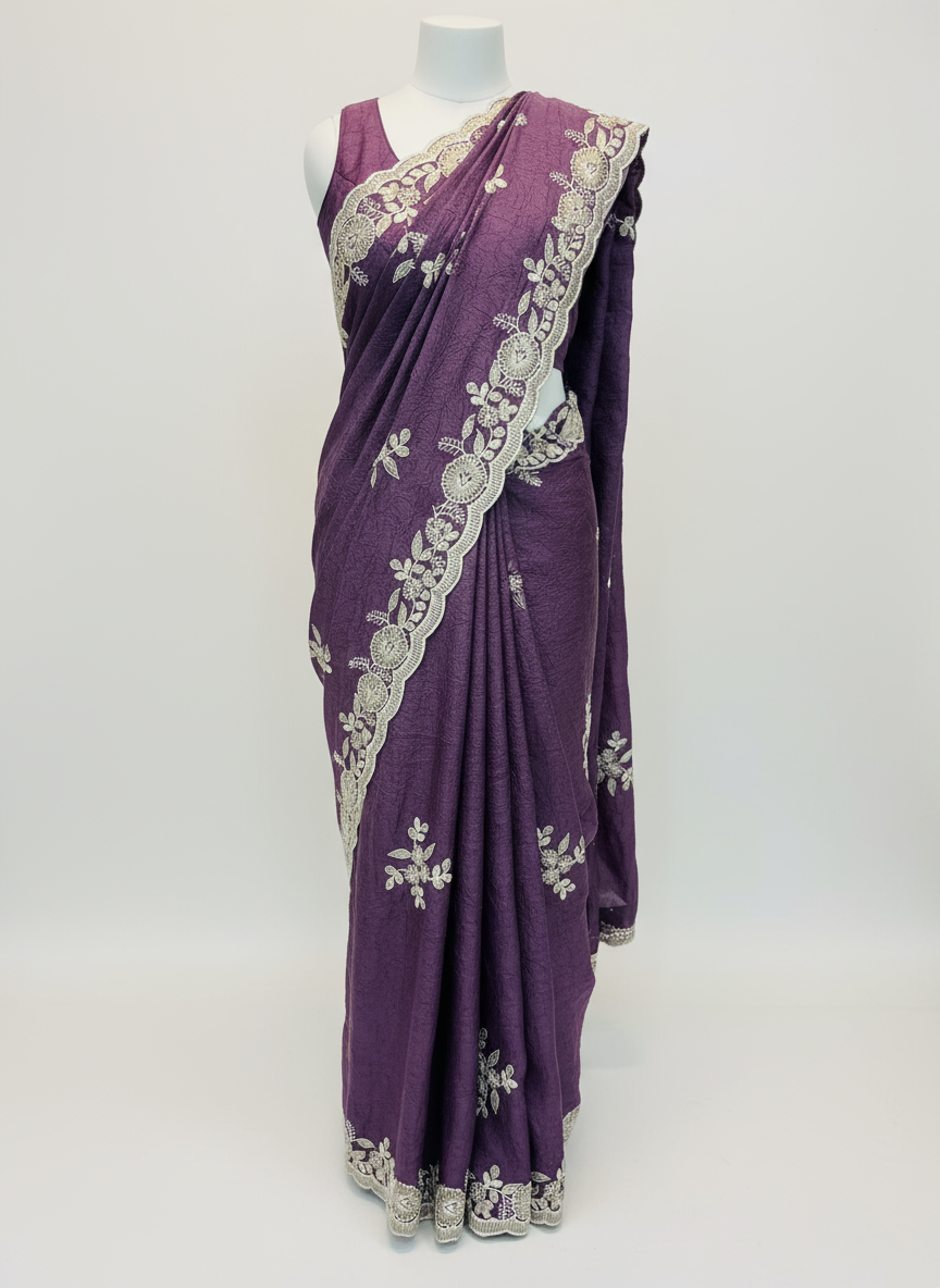 A luxurious plum purple saree featuring intricate silver floral embroidery and a scalloped border adorned with delicate sequin detailing. The fabric is paired with a matching embroidered blouse piece, displayed elegantly on a soft white fur background, showcasing its rich texture and craftsmanship.