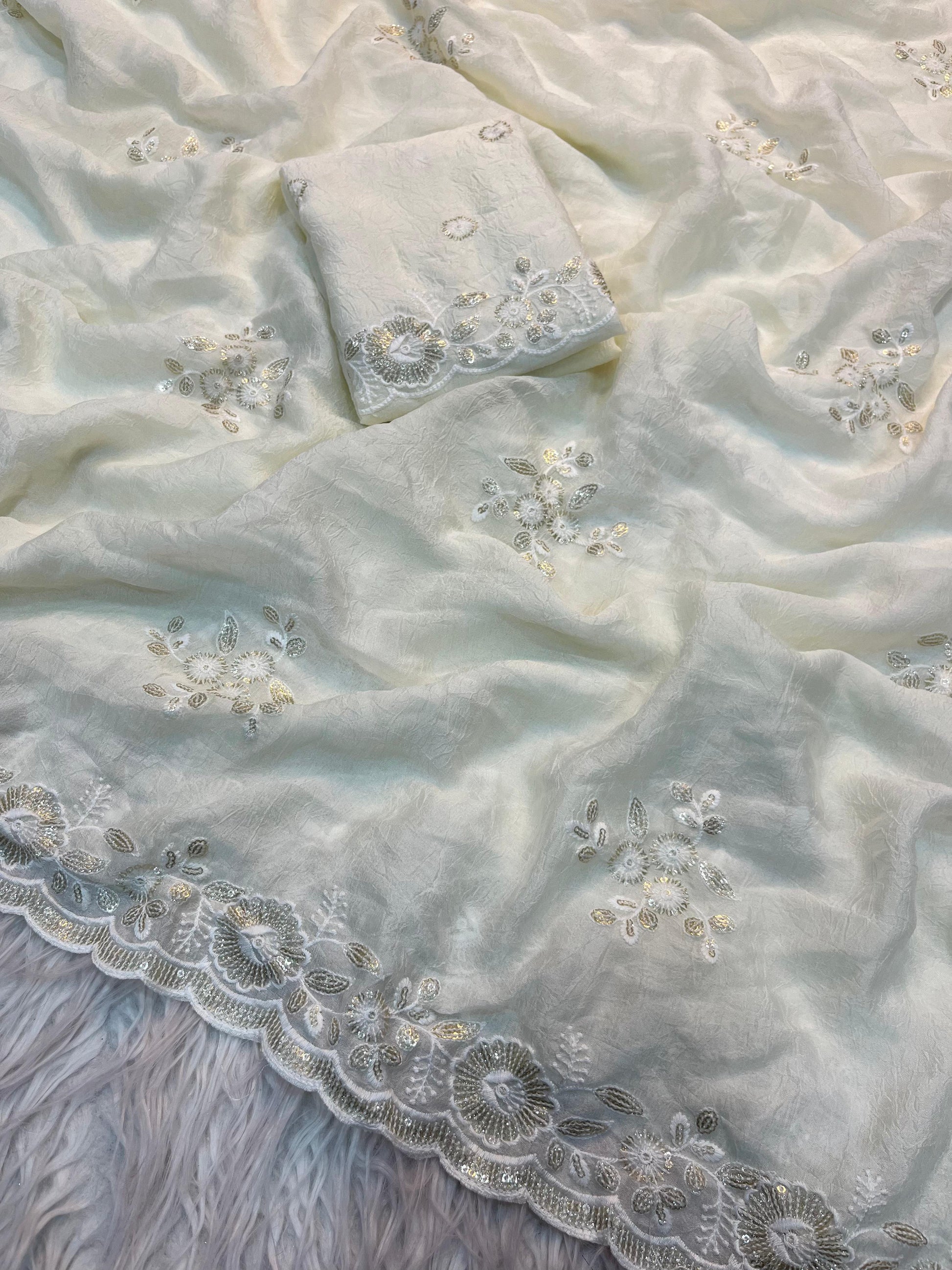 A pure white designer saree adorned with subtle silver sequin floral motifs and a beautifully scalloped embroidered border. The fabric is complemented by a matching blouse piece, creating a refined and contemporary ethnic look.