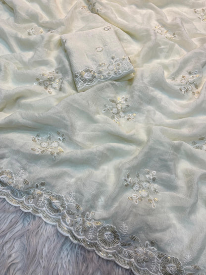 A pure white designer saree adorned with subtle silver sequin floral motifs and a beautifully scalloped embroidered border. The fabric is complemented by a matching blouse piece, creating a refined and contemporary ethnic look.