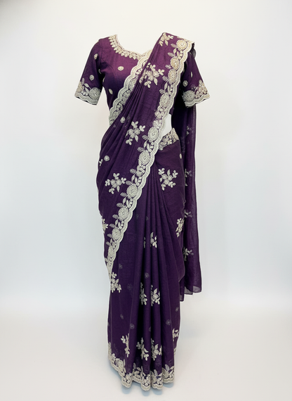 A mannequin draped in a royal purple saree featuring intricate silver zari embroidery and floral motifs throughout. The saree includes a scalloped embroidered border and matching blouse with detailed silver threadwork on the sleeves and neckline, showcasing a regal and elegant ethnic style.
