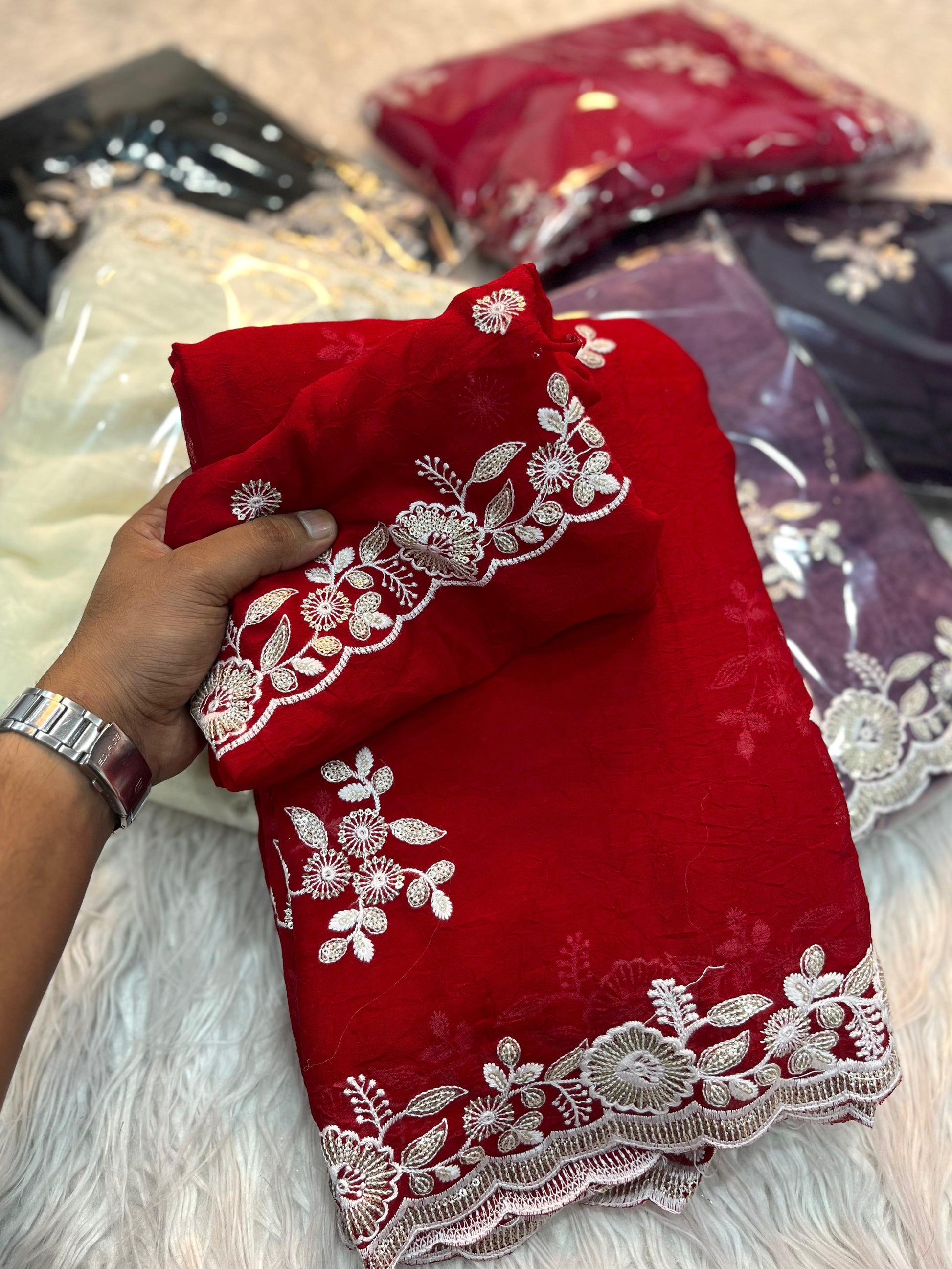 A stunning ruby red designer saree featuring delicate silver thread and sequin floral embroidery. The border is accentuated with scalloped lace detailing, paired with a matching blouse piece, creating a festive and elegant look.