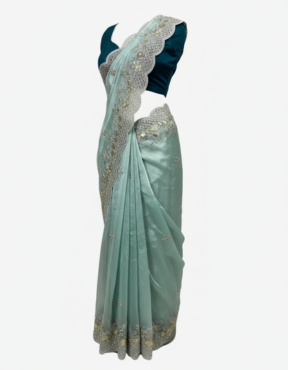 A shimmering sea green organza saree featuring delicate silver and gold sequin embroidery along a scalloped border. The saree is paired with a contrasting dark teal blouse piece, placed in a woven basket on a soft white fur background, showcasing its elegant texture and intricate craftsmanship.