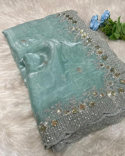 A shimmering sea green organza saree featuring delicate silver and gold sequin embroidery along a scalloped border. The saree is paired with a contrasting dark teal blouse piece, placed in a woven basket on a soft white fur background, showcasing its elegant texture and intricate craftsmanship.