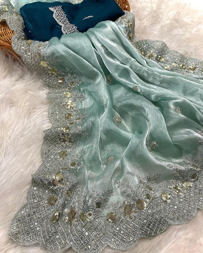 A shimmering sea green organza saree featuring delicate silver and gold sequin embroidery along a scalloped border. The saree is paired with a contrasting dark teal blouse piece, placed in a woven basket on a soft white fur background, showcasing its elegant texture and intricate craftsmanship.