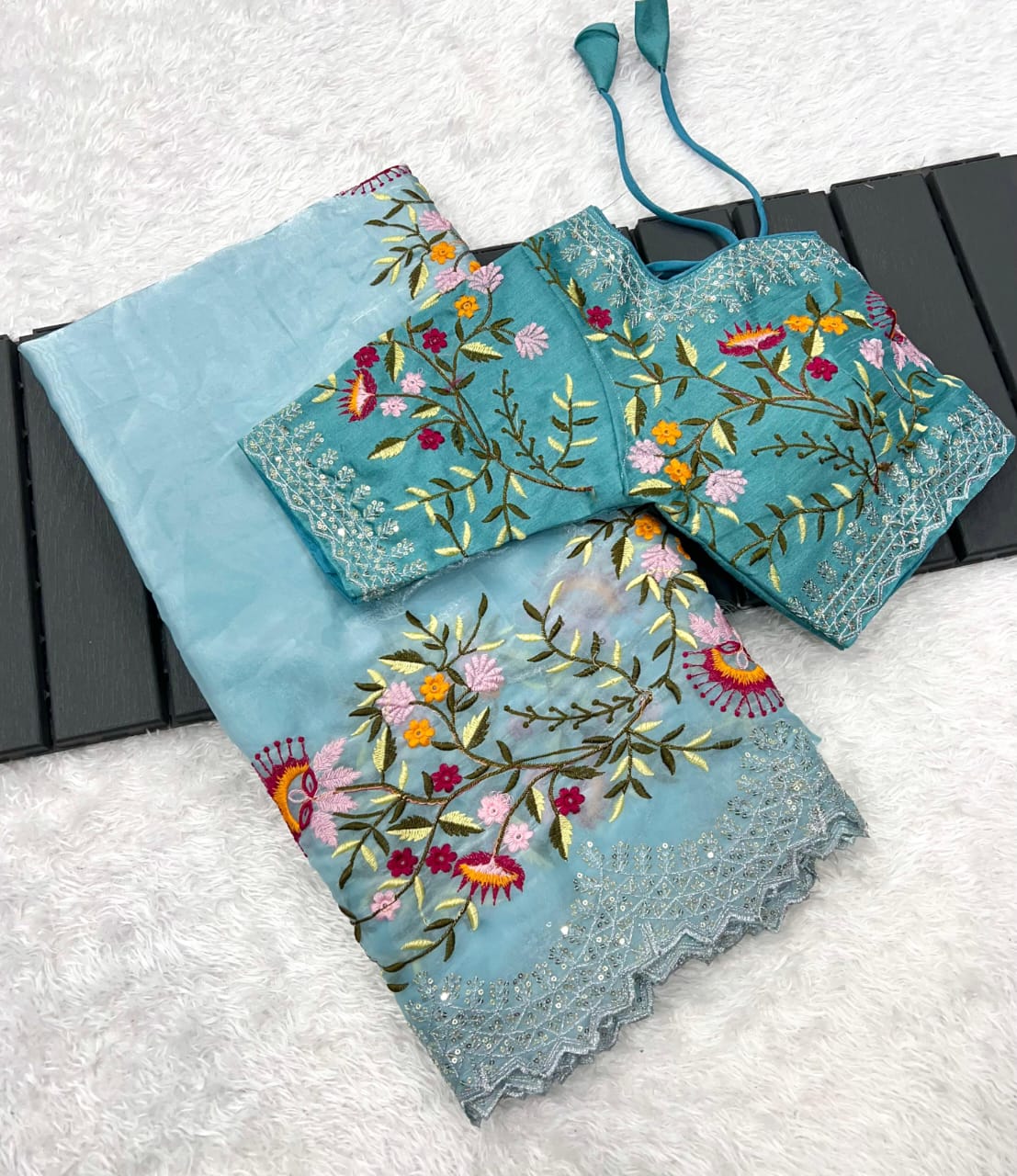 An exquisite sky blue organza saree adorned with delicate multicolor floral embroidery and a lace-trimmed border. The outfit comes with a stitched matching blouse featuring rich thread embroidery, perfect for festive and wedding occasions.