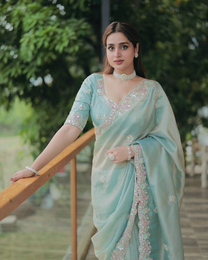 A woman wearing a space blue silk saree featuring a delicate pink and silver floral embroidered border. The saree has a soft, glossy finish paired with a matching embroidered blouse. She accessorizes with a pearl choker, earrings, and statement jewelry, posing gracefully on a balcony with greenery in the background, showcasing the saree’s elegant drape and intricate detailing.