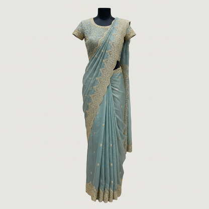 A luxurious teal blue organza saree beautifully adorned with intricate golden embroidery and heavy sequin work along the scalloped border. The saree is paired with a matching embroidered blouse piece and displayed with elegant accessories, exuding a regal and festive charm.