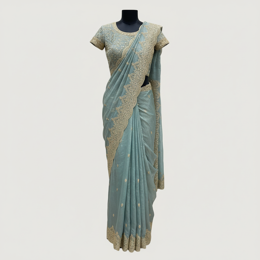 A luxurious teal blue organza saree beautifully adorned with intricate golden embroidery and heavy sequin work along the scalloped border. The saree is paired with a matching embroidered blouse piece and displayed with elegant accessories, exuding a regal and festive charm.