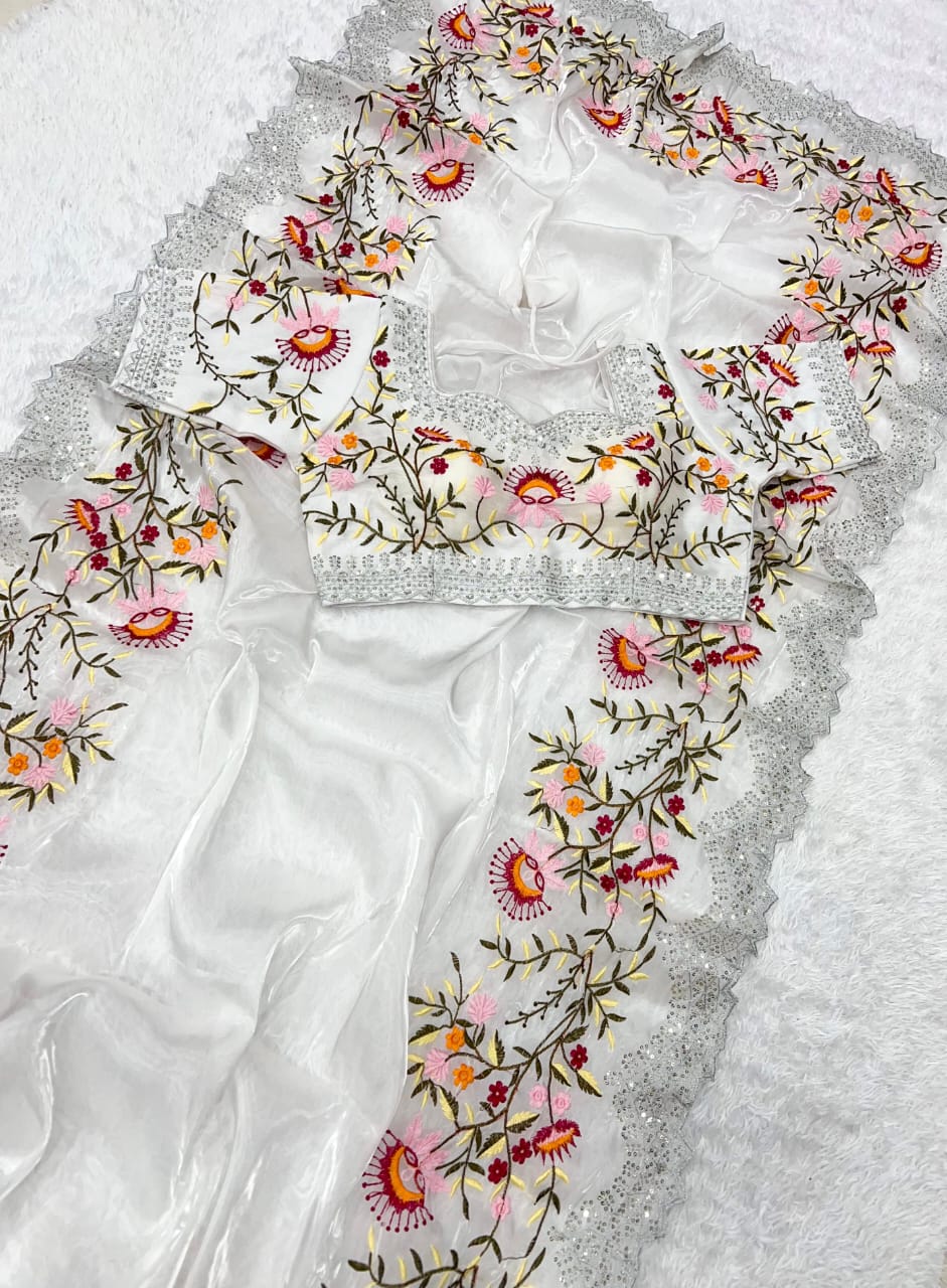 A modern white organza saree decorated with intricate multicolor thread embroidery featuring floral motifs. The stitched blouse complements the saree with identical embroidery, blending classic elegance with a contemporary touch.