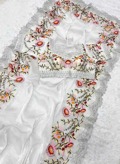 A modern white organza saree decorated with intricate multicolor thread embroidery featuring floral motifs. The stitched blouse complements the saree with identical embroidery, blending classic elegance with a contemporary touch.